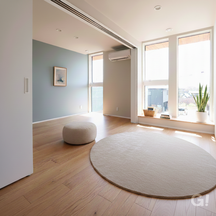 千葉県市川市の注文住宅にある、開放的で明るい2階フリースペース。ブルーグレーのアクセントウォールに小さなアートを飾り、床には円形のラグとベージュのオットマンを配置した北欧スタイルの空間。大きな窓からは自然光が降り注ぎ、窓辺の造作カウンターにはサンセベリアの